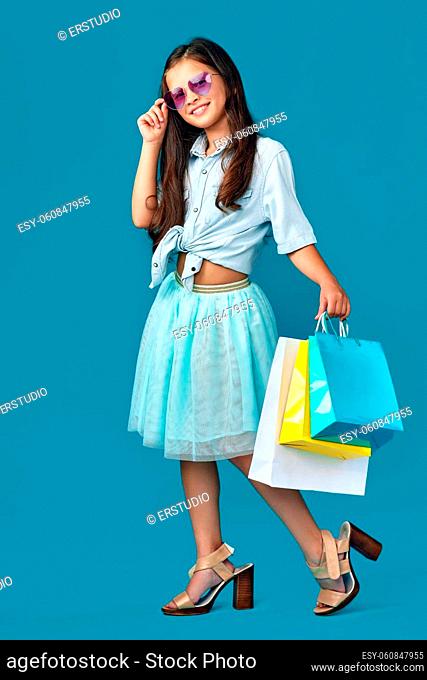 happy child girl in sunglasses holds many shopping bags with purchases isolated on blue background