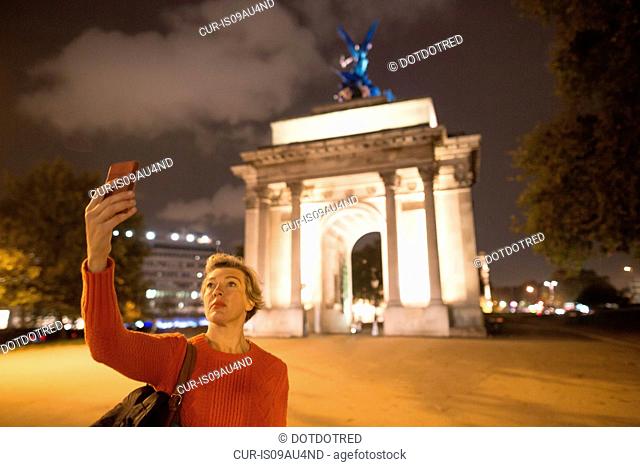 Mature female tourist taking smartphone selfie of Wellington Arch at night, London, UK