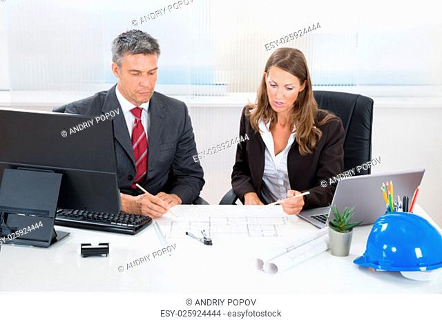Two Architects Together Discussing Blueprint At Desk In Office