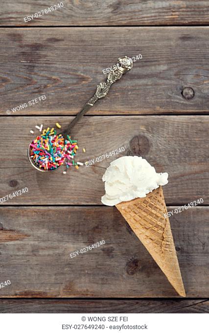 Top view single scoop scoop vanilla ice cream in waffle cone with spoon of color rice on wood background