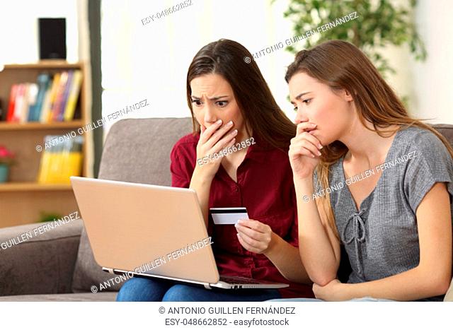 Two worried friends having problems buying on line with credit card and a laptop sitting on a couch in the living room at home
