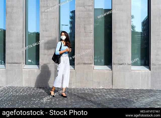 Businesswoman in face mask standing on footpath by building during coronavirus crisis