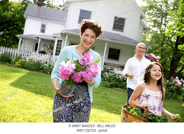 A summer family gathering at a farm. Parents and children walking across the lawn carrying flowers, fresh picked vegetables and fruits