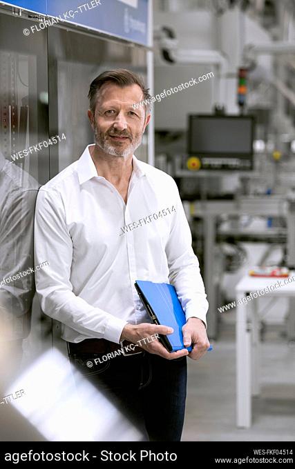 Mature businessman holding file while leaning on glass in factory