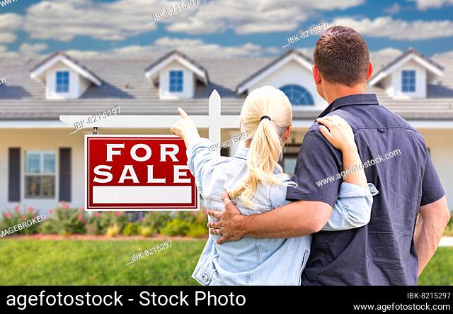Young adult couple facing and pointing to front of for sale real estate sign and house