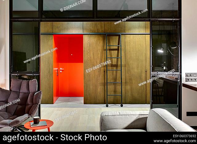 Luminous hall in a loft style with a red entrance door, wooden wardrobes and lockers, ladder with wheels, armchair, table with book and cup, sofa