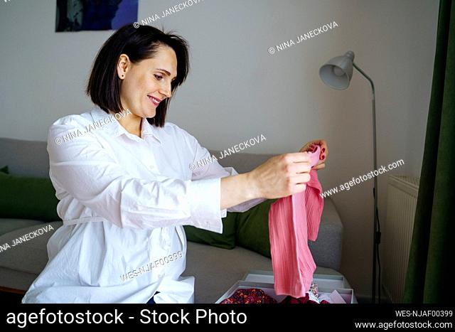 Happy pregnant woman looking at baby clothes at home