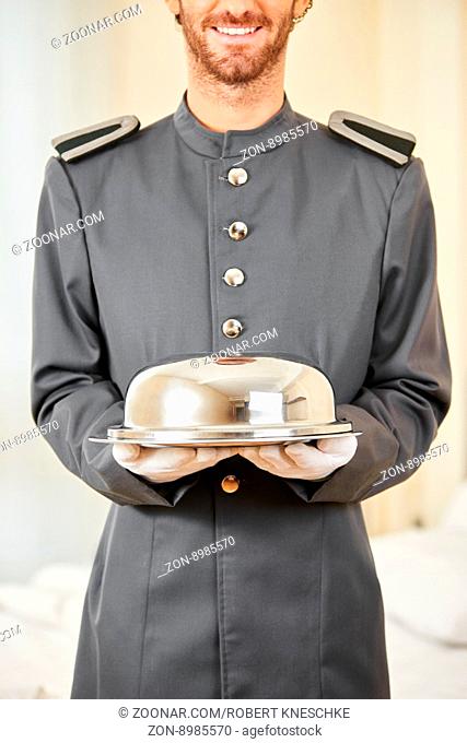 Freundlicher Page serviert das Essen mit Speiseglocke auf einem Tablett im Hotel
