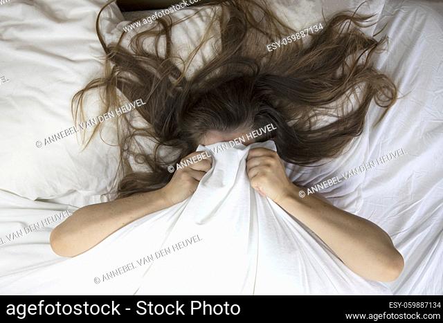 Young woman lying in bed suffering, tired woman covering face with hands, can't sleep feels exhaustand on white sheets in bedroom closeup