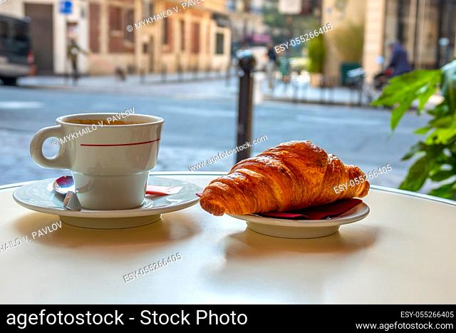 Table of street cafe in Paris in the morning. Cup of coffee and croissant close-up. Background in defocus