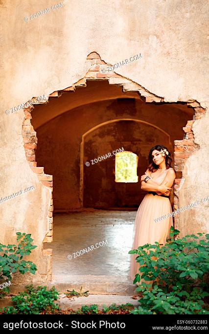 Lovely bride in a beautiful pastel dress with her arms crossed on her chest is pensively standing on the ruins of a castle. High quality photo