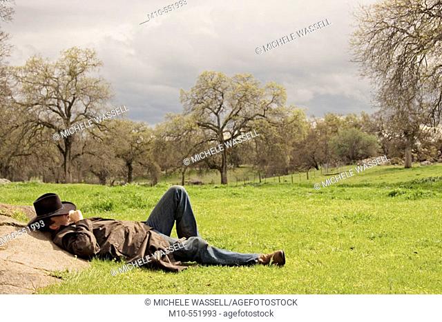 Cowboy in western attire laying down up against a rock during the break of a storm in a beautiful countryside landscape