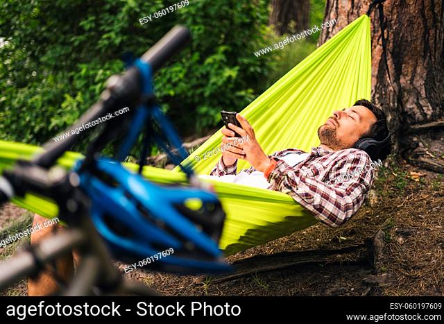 Man on bicycle trip at camping by lake is relaxing in green hammock while listening to music. Active recreation theme in nature