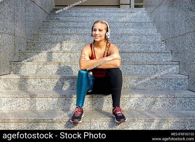 Smiling sportswoman with prosthetic leg wearing headphones sitting with arms crossed on staircase