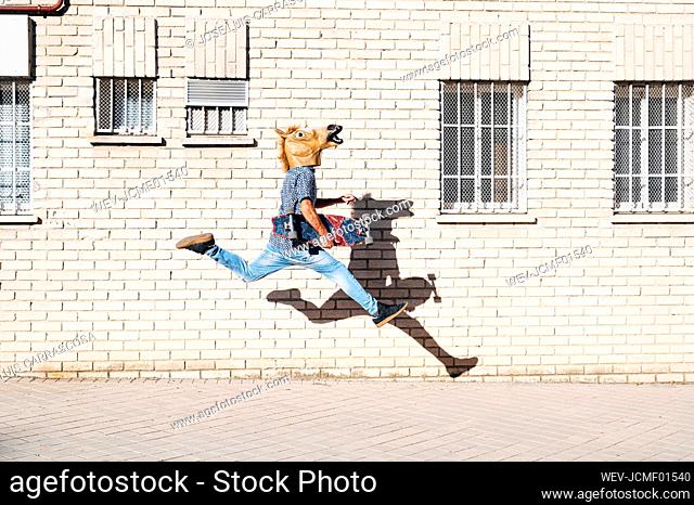 Man wearing horse mask holding skateboard while jumping on footpath by building in city during sunny day