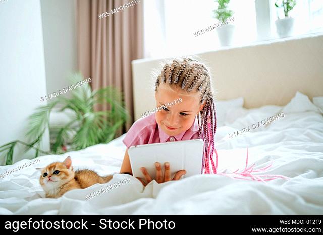 Girl holding tablet PC lying by ginger kitten on bed