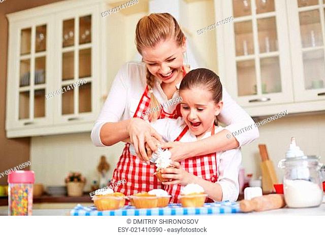 Young woman showing her daughter how to decorate baked muffins with whipped cream