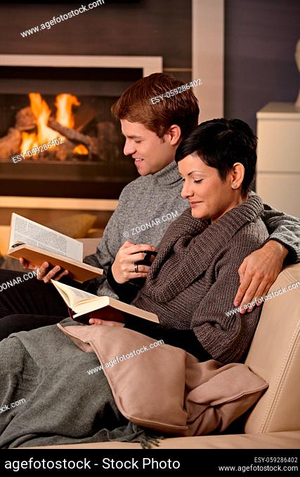 Young couple hugging on sofa in front of fireplace at home, reading books, smiling