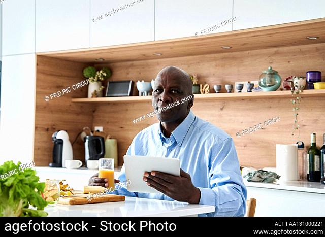 Senior African-American man sitting in kitchen eating fruit for breakfast while checking messages on digital tablet