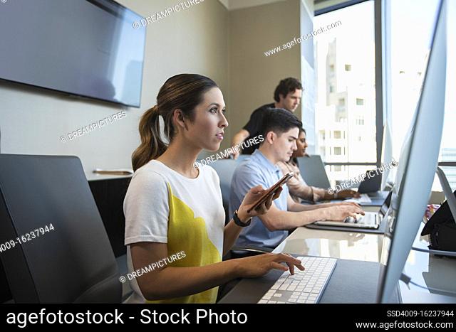 Young woman using computer in office typing on keyboard, and checking mobile phone with co-workers working in background