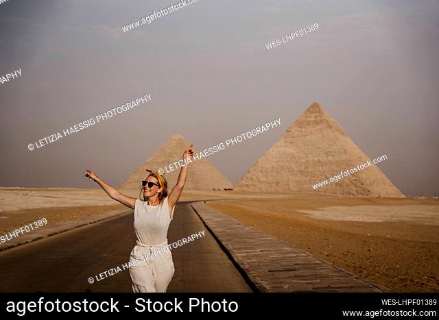 Female tourist standing with raised arms in middle of road leading to Giza Pyramids