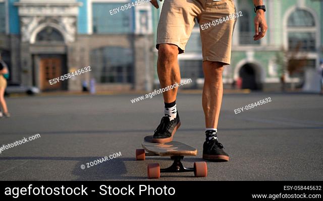 The skater's legs begin to move on a skateboard in the fresh air. One leg stands on the asphalt, the other on the longboard. Sunny blurred background