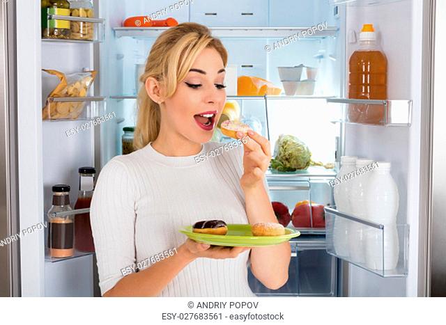Young Woman Standing Near Refrigerator Eating Donut From Plate