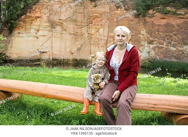 Happy Kid Girl Sitting with Mother on Wooden Tree Trunk Bench by Sandstone Outcrop