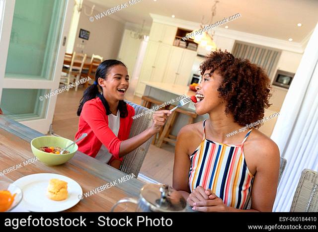 Happy african american daughter feeding mother sitting at table during breakfast and smiling