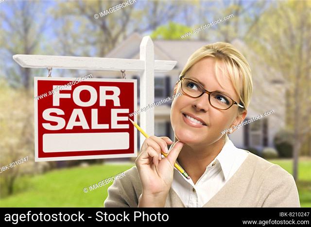 Attractive young adult woman with pencil in front of for sale real estate sign and house