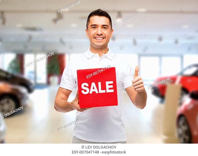shopping, discount, consumerism, gesture and people concept - smiling man with red sale sigh showing thumbs up over auto show background