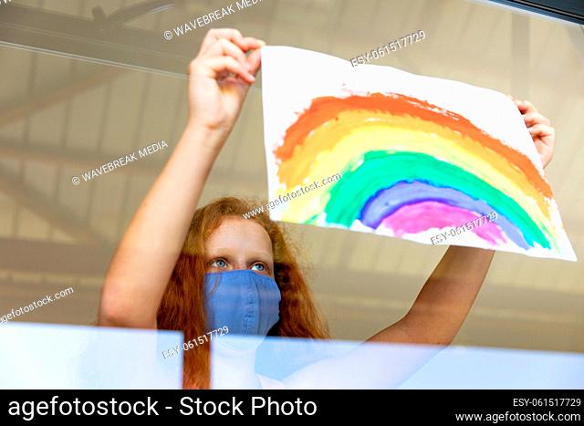 Girl wearing face mask sticking rainbow painting to the window