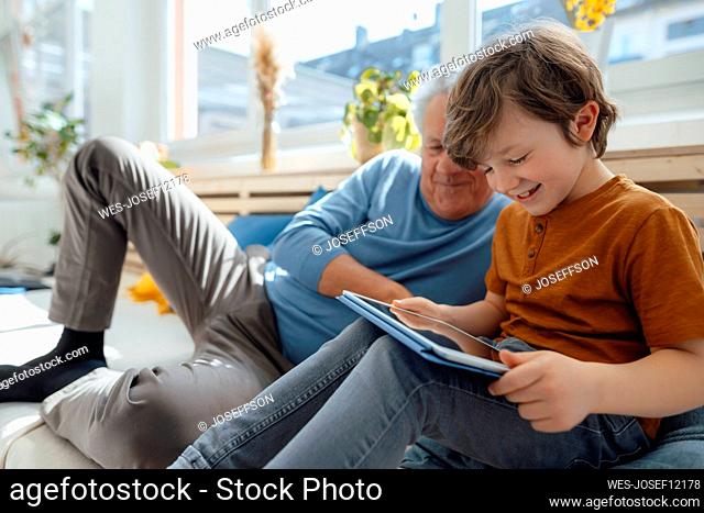 Happy grandson using tablet PC by grandfather in living room
