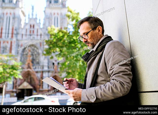 Businessman holding clipboard standing by wall in city