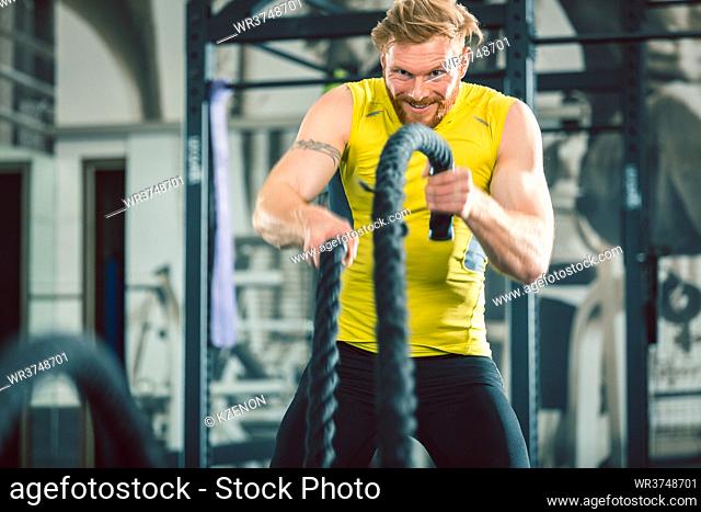 Full length low angle view of a handsome bodybuilder exercising with heavy battle ropes during intense functional training workout at the gym