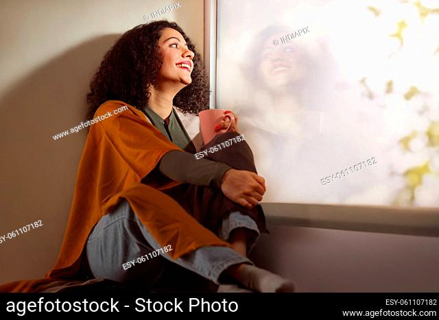 Happy young adult woman holding cup of coffee near window with reflection in modern living room