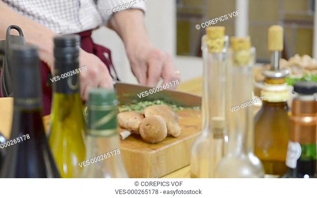 Defocussed shot of a chef's hands, cutting spices and herbs on a cutting board, seen through various bottles of vinaigrette and oil