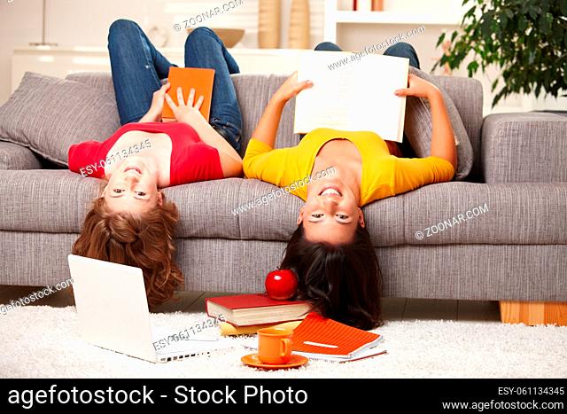 Happy teen girls sitting upside down on sofa smiling looking at camera holding books