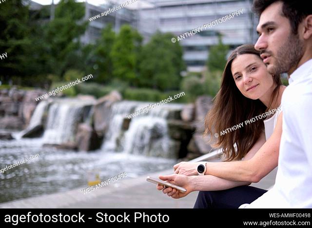 Smiling businesswoman with colleague sitting by pond at office park