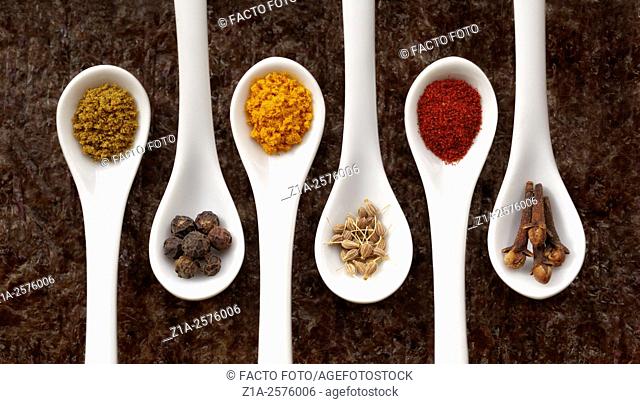 Five white spoons with assorted spices on a dark background
