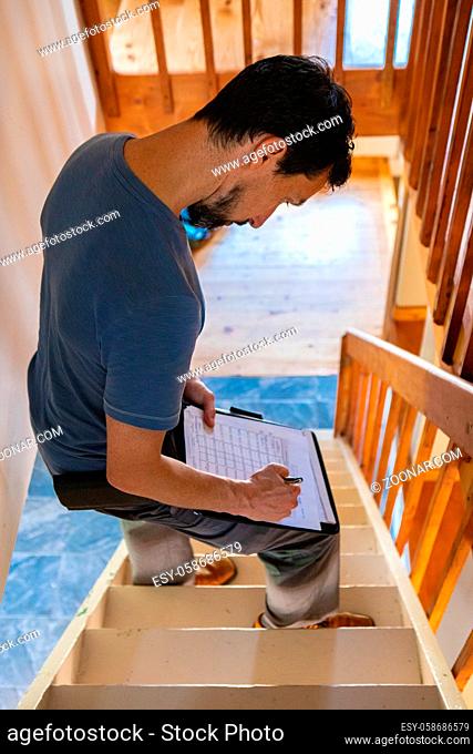 A high angled view of a home inspector standing on stairs inside a family home, taking notes during an indoor environmental quality IEQ assessment