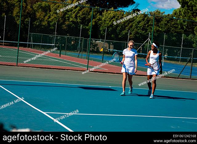 Smiling multiracial female players talking while walking at tennis court on sunny day