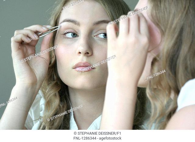 Reflection of young woman using eyebrow tweezer in mirror