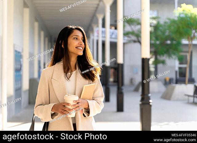Smiling young woman with book and takeaway coffee on the go in the city