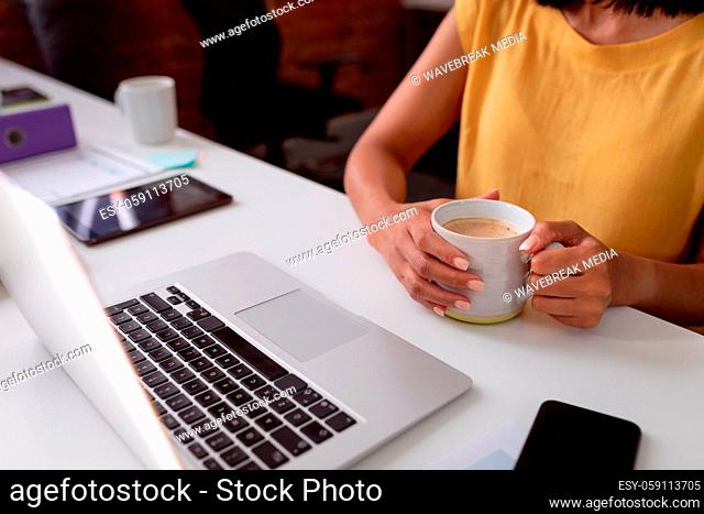 Midsection of caucasian businesswoman sitting at desk using laptop and holding cup of coffee