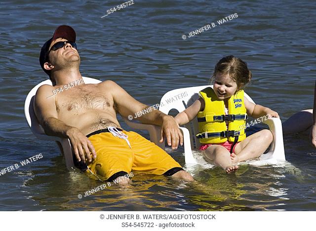A man, 20-30, and a little girl cool off sitting in chairs in a lake