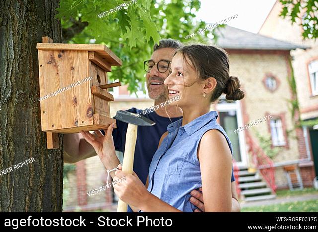 Smiling girl with father hanging nest box on tree trunk in back yard
