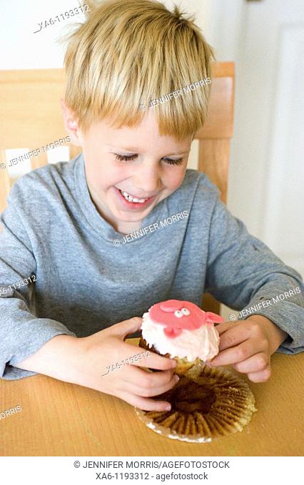 A young blond haired boy smiles as he unwraps and prepares to eat a cupcake with a pink pig on it