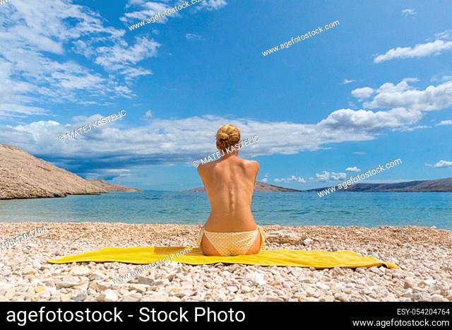 Rear view of relaxed sexy young caucasian woman sitting on yellow towel, sunbathing topless alone on romote pabble beach on Pag island, Croatia, Mediterranean