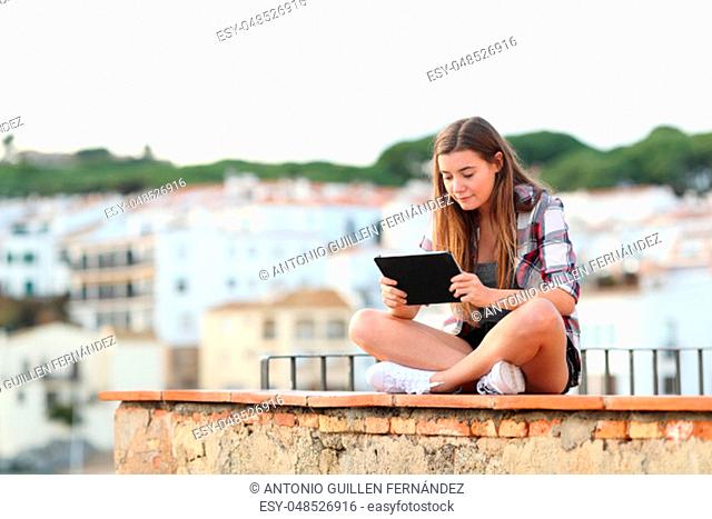 Full body portrait of a serious girl using a tablet sitting on a ledge in a town